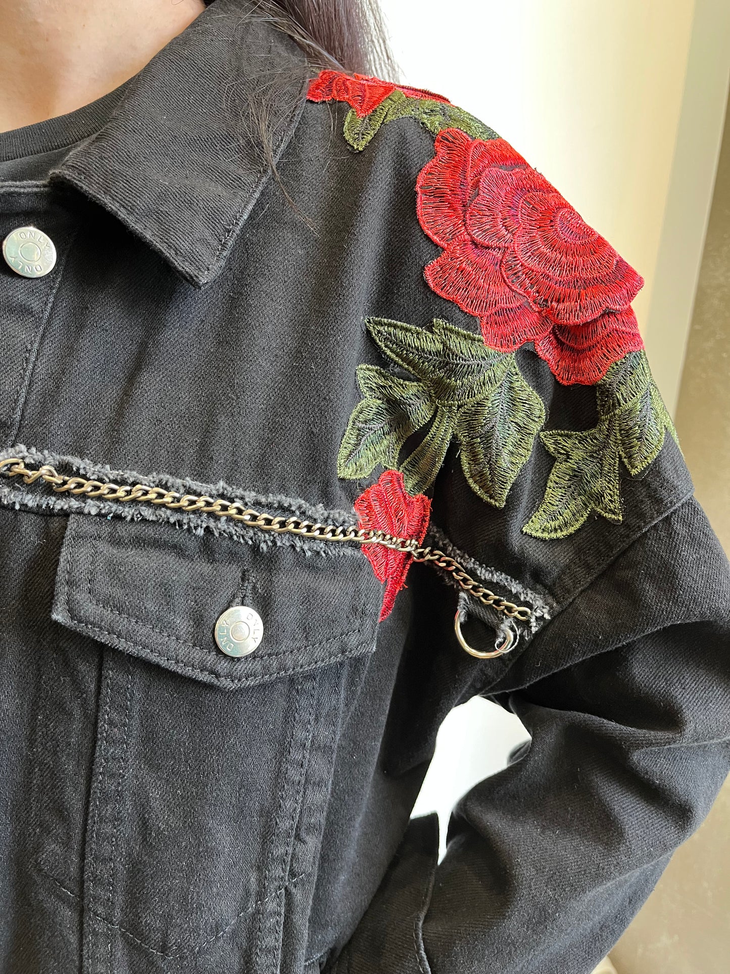 Detalle de chaqueta negra con bordados de rosas rojas y verdes en el hombro y cadena decorativa plateada en el pecho.
