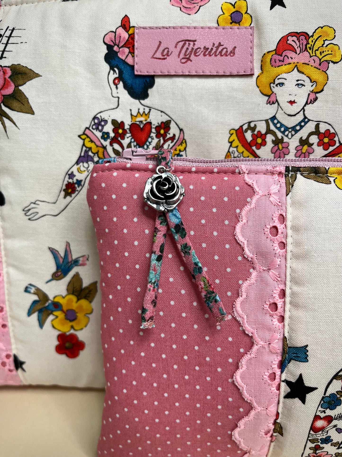 Detalle de un neceser de la colección ‘Tattoo Ladies’ con diseño que combina tela rosa de lunares blancos, encaje rosa y un tirador decorativo en la cremallera con un charm metálico en forma de rosa. De fondo se observa un estampado de mujeres tatuadas y motivos vintage con flores y golondrinas, junto a una etiqueta de tela con el nombre ‘La Tijeritas’.
