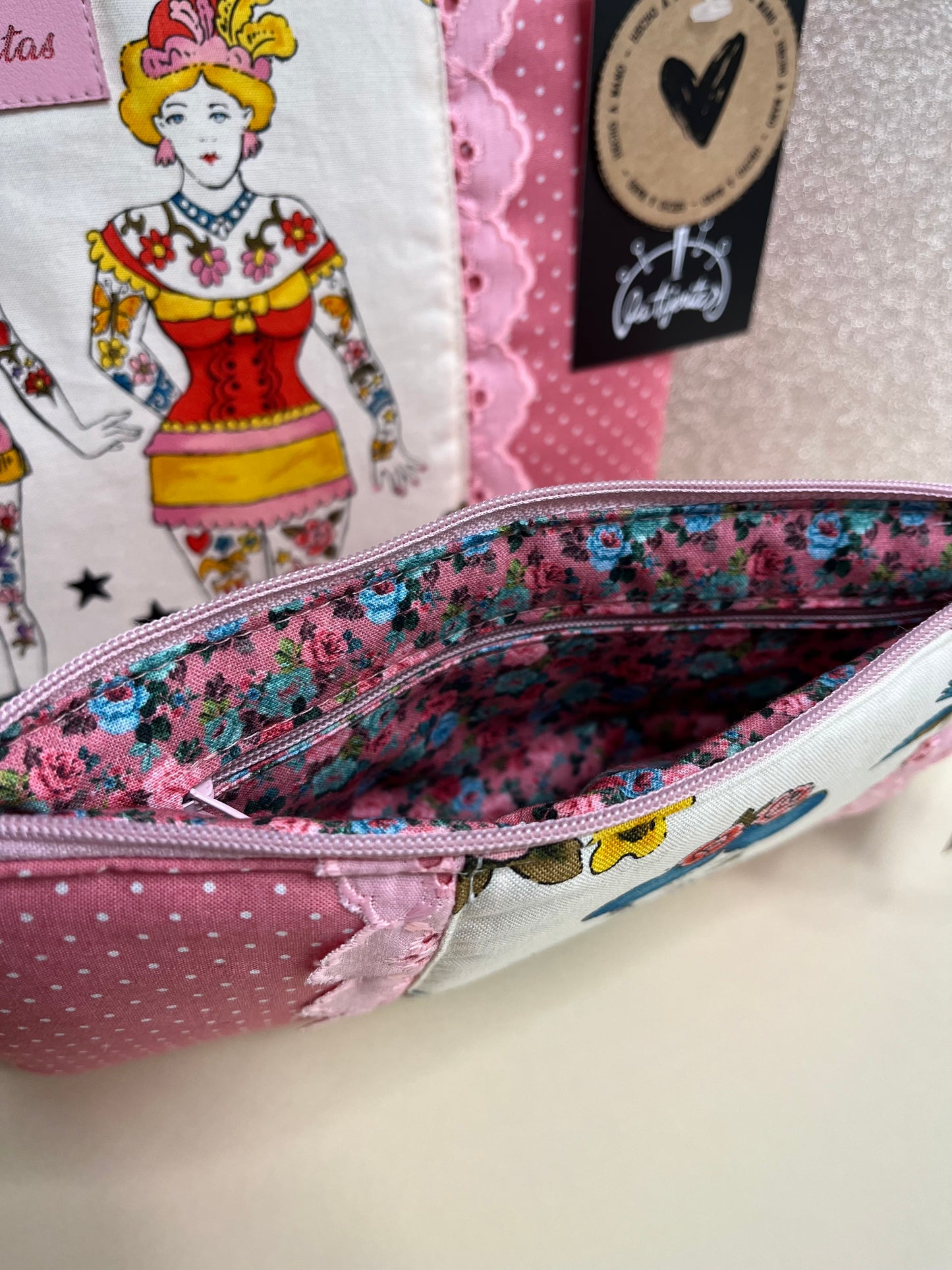 Vista del interior de un neceser de la colección ‘Tattoo Ladies’ con diseño exterior de tela rosa con lunares blancos, encaje rosa y estampado de mujeres tatuadas. El forro interior es floral en tonos rosas y azules e incluye un compartimento adicional con cremallera.