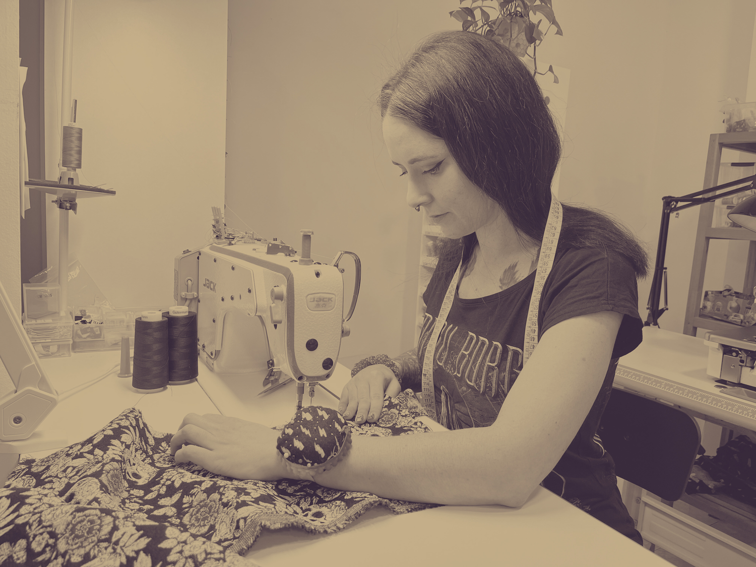 Fotografía en blanco y negro de una mujer trabajando con una máquina de coser industrial. Lleva una cinta métrica alrededor del cuello y un cojín para alfileres en la muñeca mientras manipula una tela con estampado floral. En la mesa se observan hilos y herramientas de costura. Al fondo, se ven plantas y estanterías con materiales de trabajo.