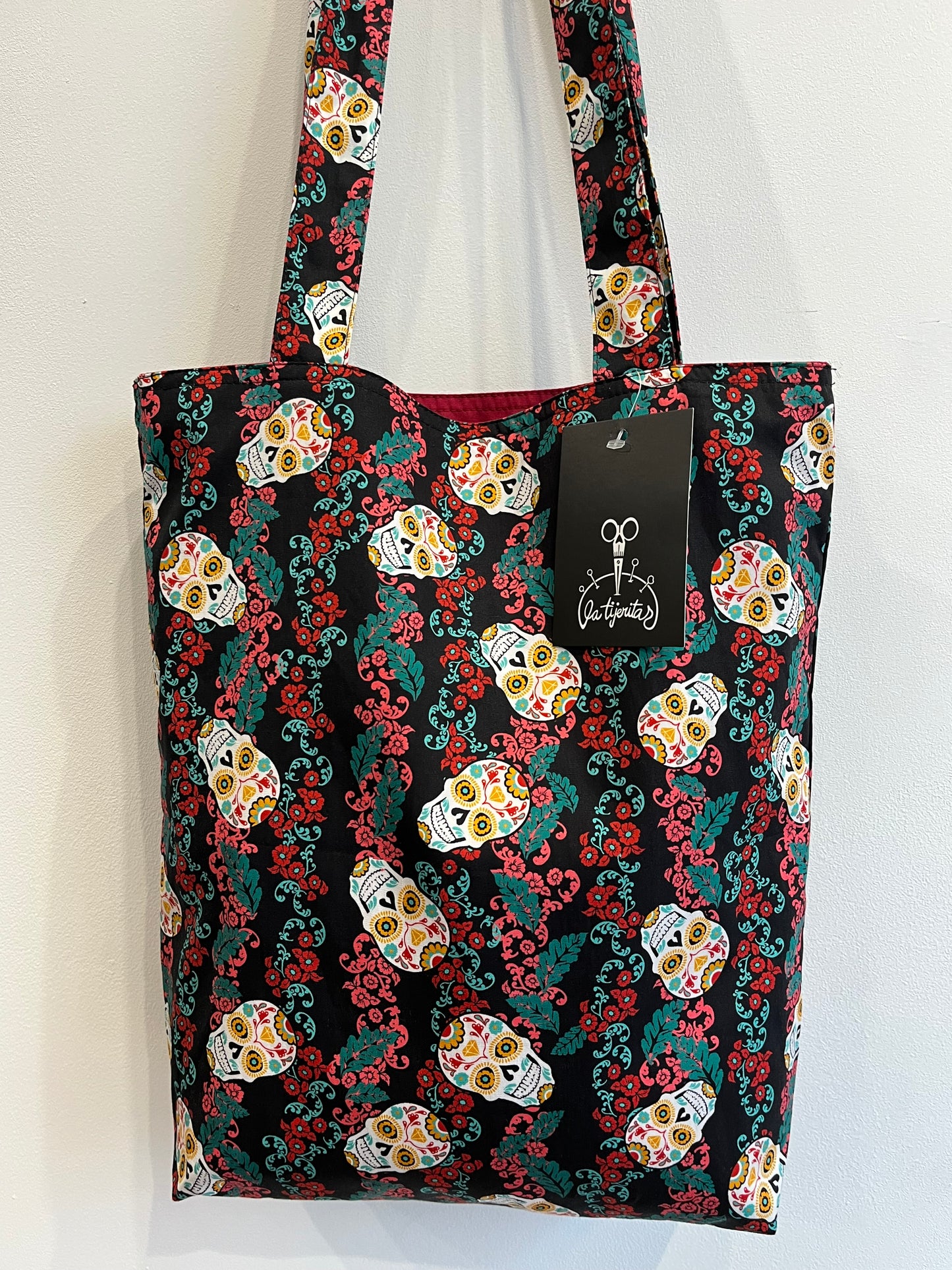 Bolso de tela negra con estampado de calaveras mexicanas coloridas y detalles florales en rojo y verde.