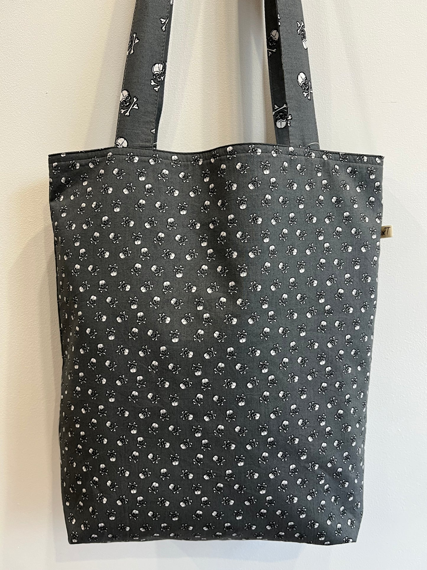 Bolso de tela gris oscuro con estampado de pequeñas calaveras blancas y detalles de huesos cruzados.