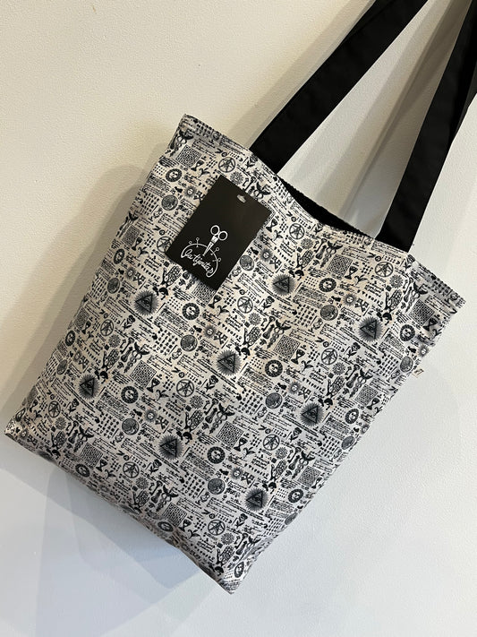 Bolso de tela blanca con estampado esotérico en negro, símbolos místicos y asas negras, con etiqueta de “La Tijeritas”.