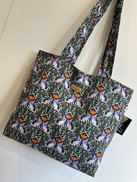 Bolso de tela con estampado de gatos lilas con sombreros de bruja y murciélagos naranjas, acompañado de detalles vegetales.