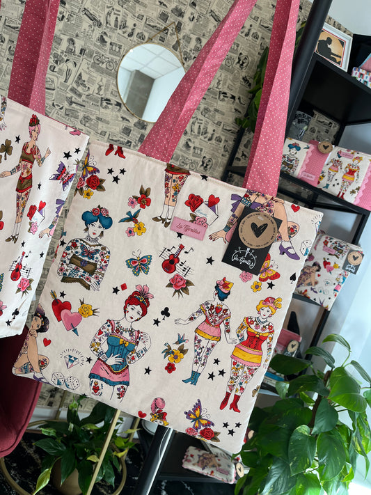 Bolso de la colección ‘Tattoo Ladies’ con estampado colorido que incluye mujeres tatuadas, flores, mariposas y corazones. Las asas son rosas con lunares blancos, y el bolso lleva una etiqueta de tela con el nombre ‘La Tijeritas’ y una etiqueta colgante que dice ‘Hecho a mano’. De fondo se observa una decoración de estilo vintage con plantas y estanterías que muestran más productos.