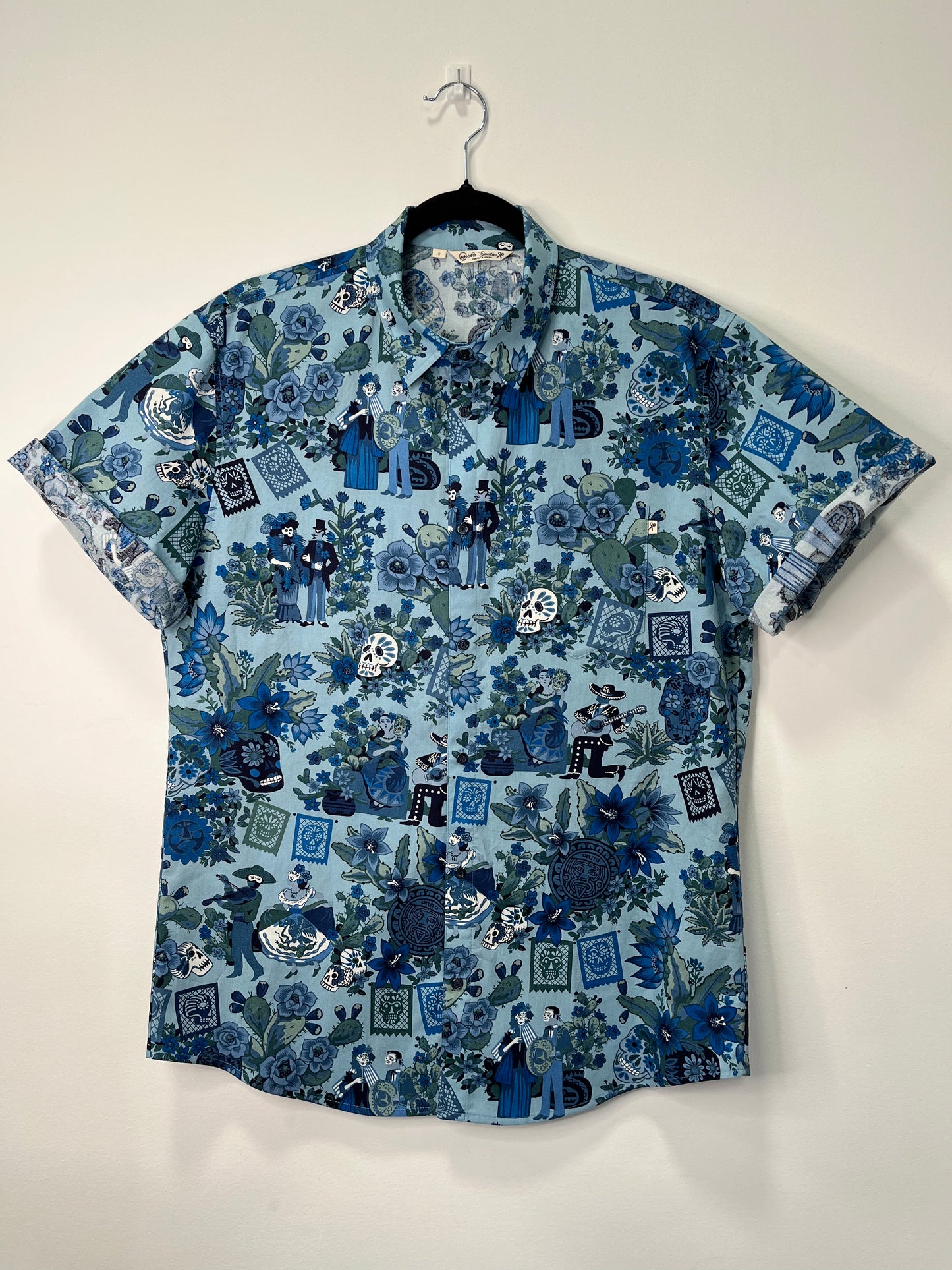 Camisa de manga corta colgada en una percha, con un estampado en tonos azules y verdes inspirado en el Día de los Muertos. El diseño incluye calaveras, flores y figuras decorativas sobre un fondo azul claro.