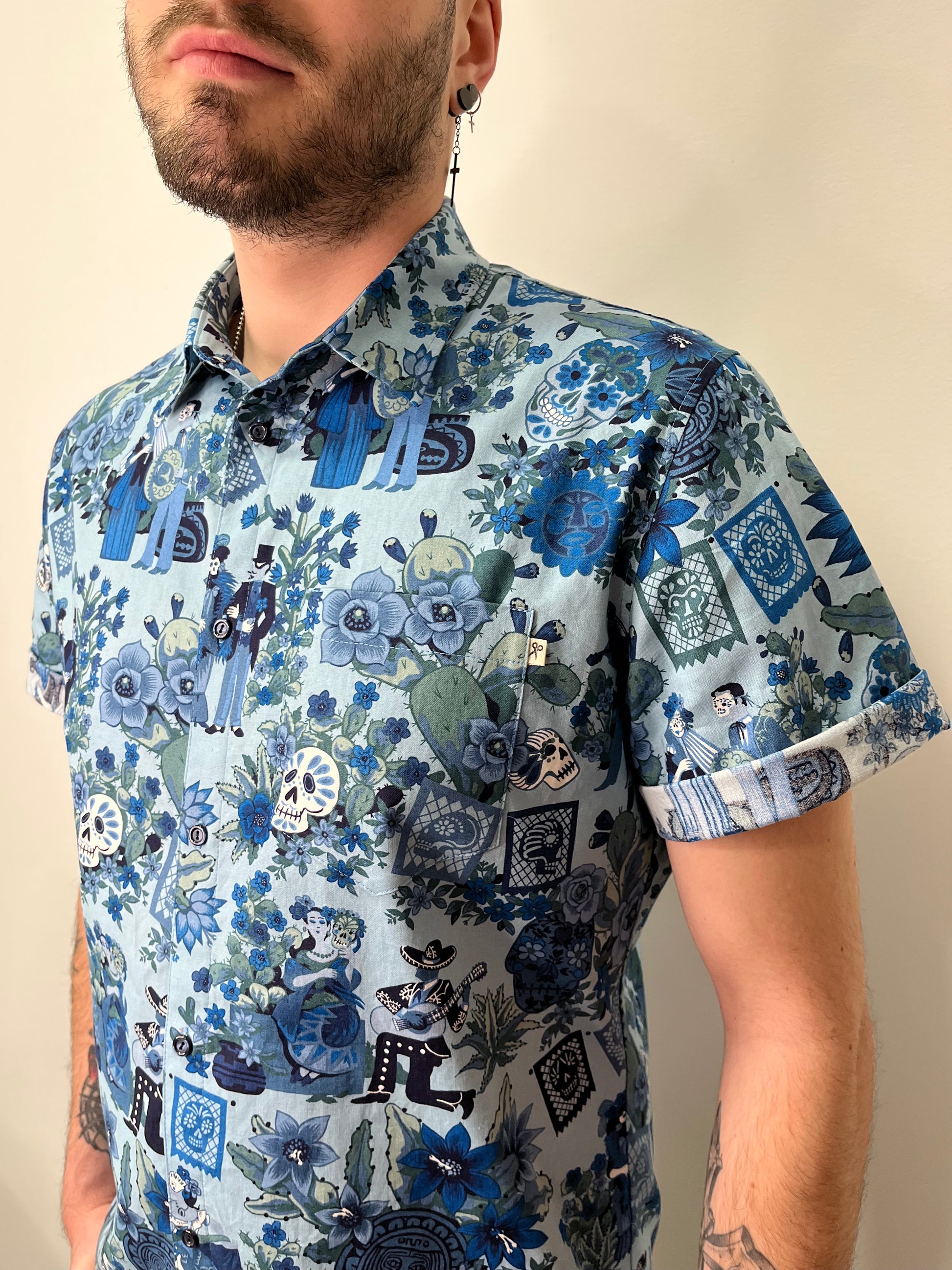 Persona vistiendo una camisa de manga corta con estampado inspirado en el Día de los Muertos, en tonos azules y verdes. El diseño incluye calaveras, flores y figuras decorativas. La foto muestra detalles del estampado y accesorios como un pendiente de cruz