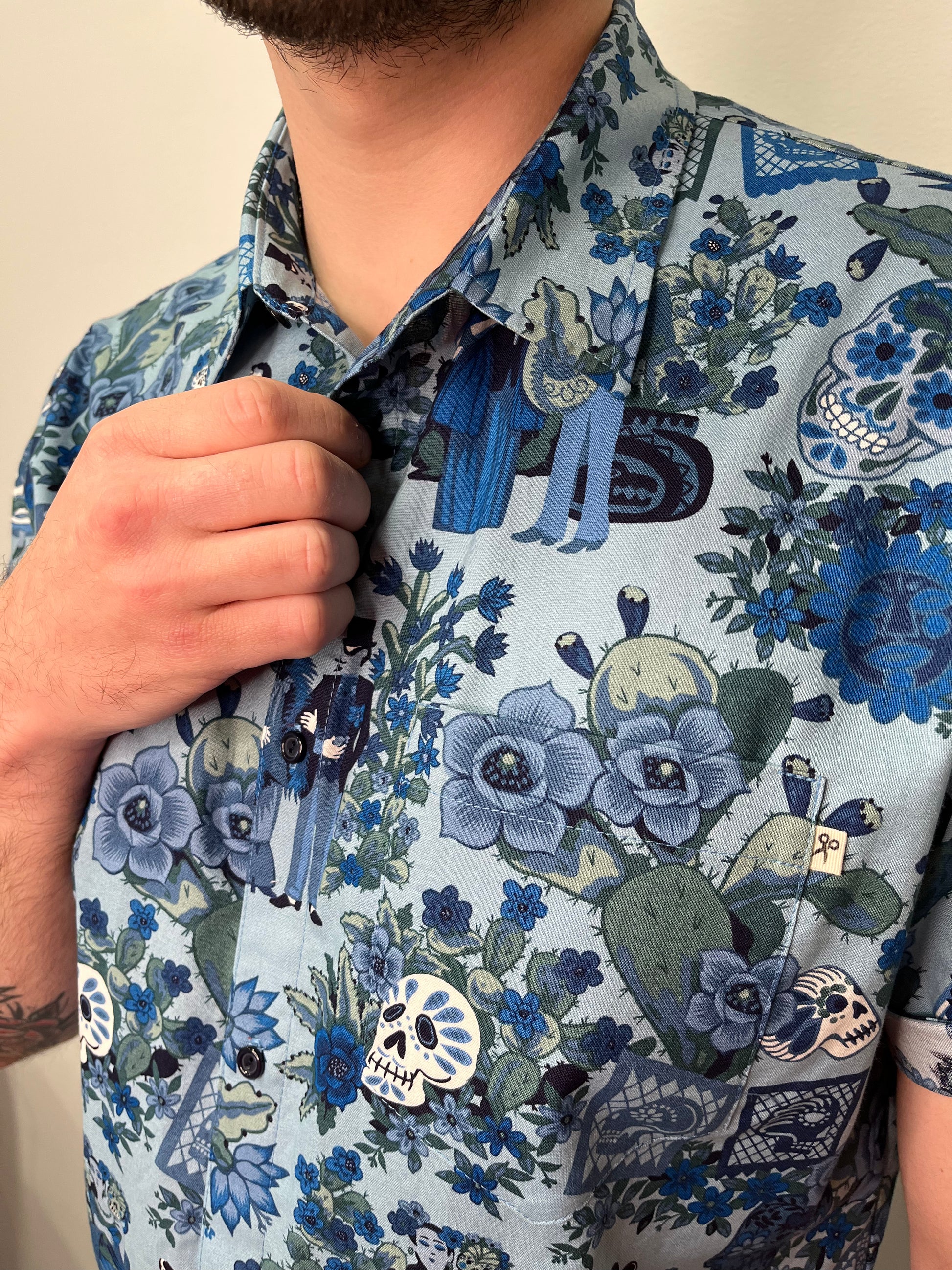 Camisa con estampado detallado de estilo Día de los Muertos en tonos azules y verdes. Incluye motivos florales, calaveras y figuras decorativas. Una persona la lleva puesta, sujetando el cuello de la camisa con la mano.