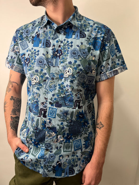 Camisa de manga corta con estampado en tonos azules y verdes, inspirada en el Día de los Muertos. Incluye diseños de calaveras, flores y figuras decorativas. La persona que la lleva tiene las manos en los bolsillos y muestra tatuajes en los brazos.