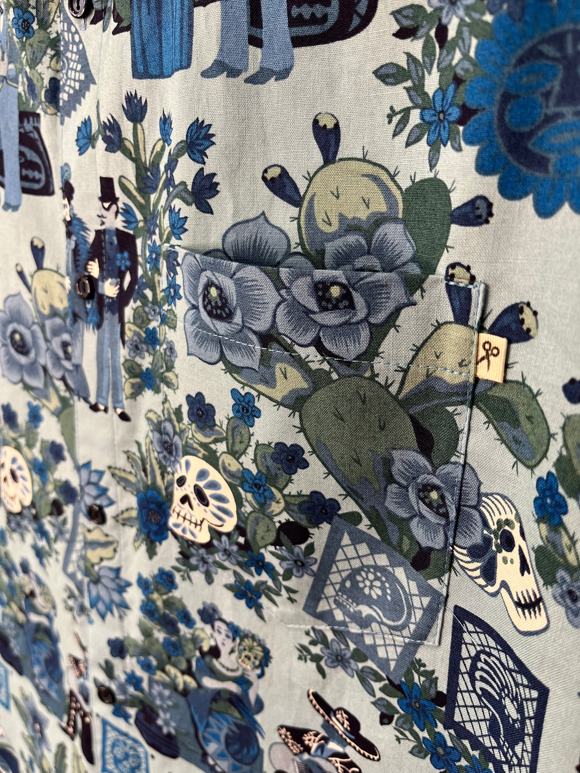 Plano detallado del estampado de una camisa inspirada en el Día de los Muertos. Se observan flores, cactus, calaveras y figuras decorativas en tonos azules y verdes, junto a un bolsillo con una pequeña etiqueta de tijeras.