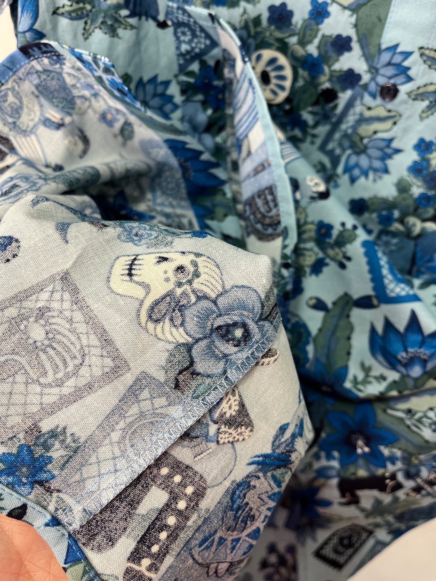 Detalle cercano del tejido de una camisa con estampado inspirado en el Día de los Muertos. Se aprecian calaveras, flores y patrones decorativos en tonos azules y verdes, con textura visible en la costura.