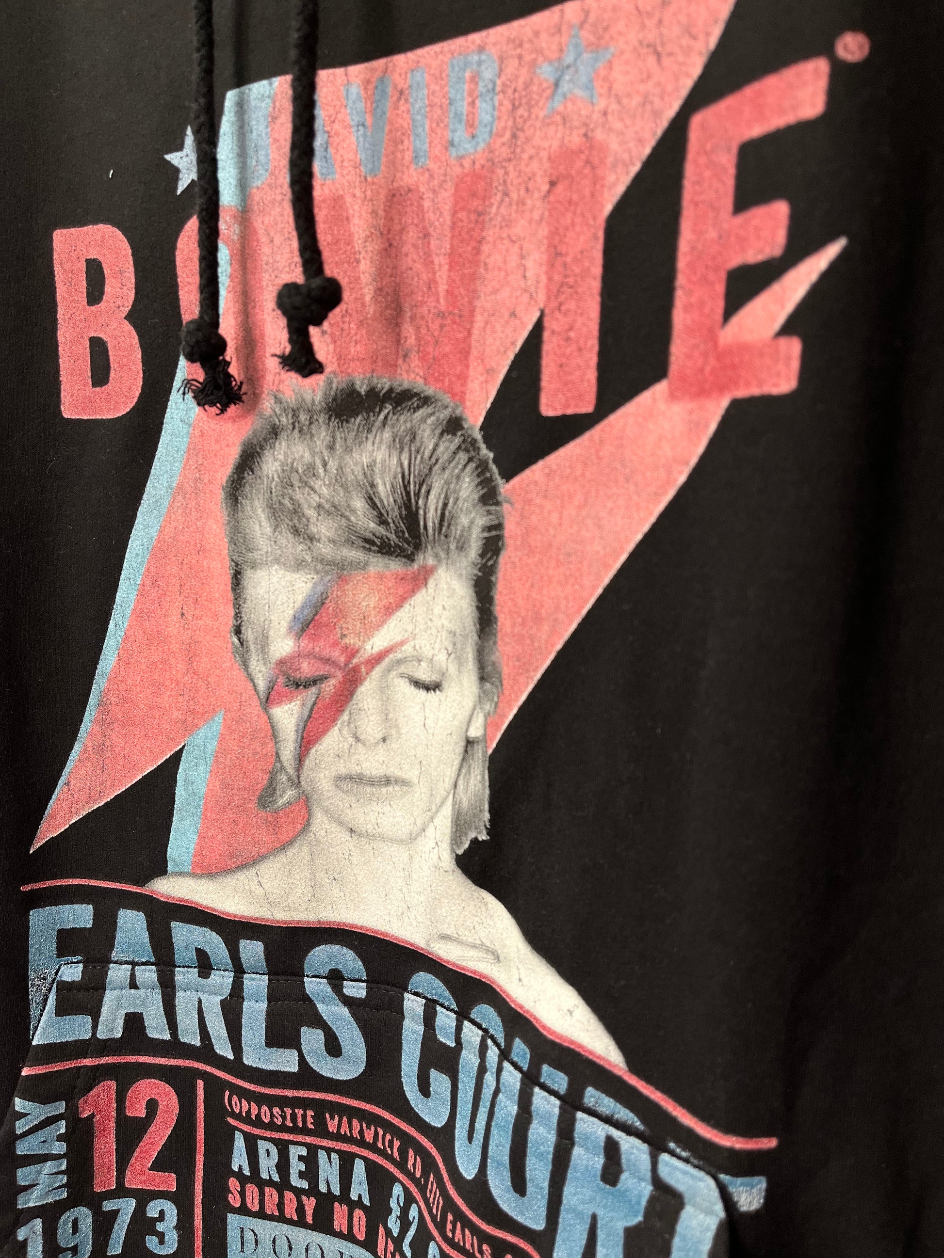 Primer plano de la serigrafía en una sudadera negra que muestra un diseño colorido inspirado en David Bowie. Incluye su icónico rayo rojo y azul en el rostro, junto a texto en letras grandes que dice ‘David Bowie’ y detalles adicionales como ‘Earls Court’ y la fecha ‘May 12, 1973’ en colores rojos y azules.