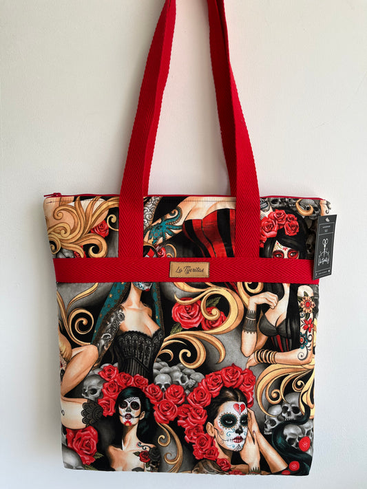 Bolso con estampado de chicas con cara de catrina muy sexys. Tatuadas, con flores rojas en la cabeza y con vestidos de encaje. El estampado es mayoritariamente gris, nergro y rojo con detalles azules. Tiene tiras a contraste en color rojo.