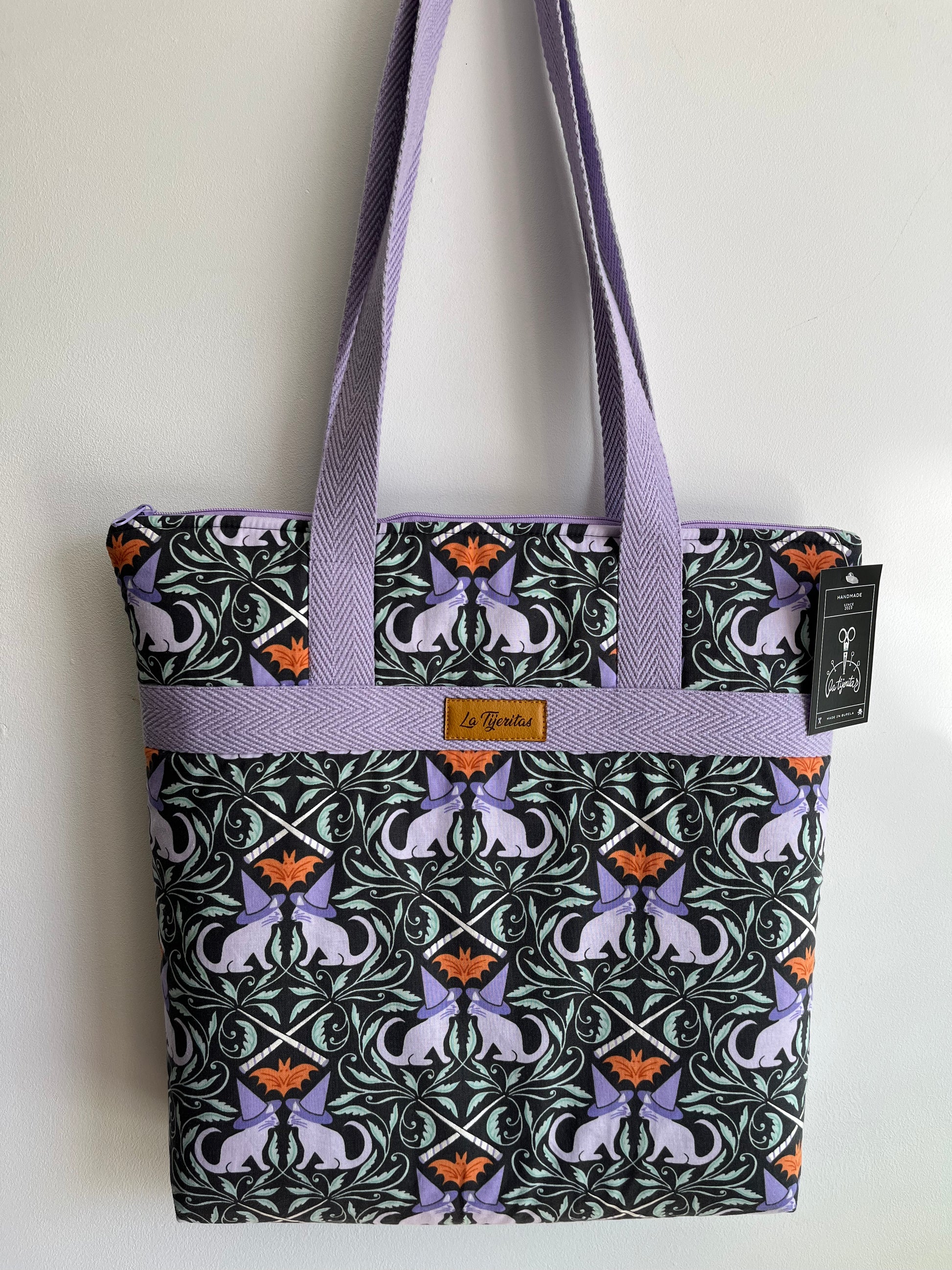 Bolso con estampado de gatos con gorritos de bruja. Fondo gris oscuro, los gatos son lila y se pueden ver murciélagos naranja,