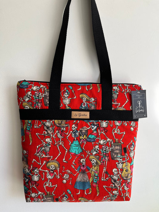 Tote bag acolchada de color rojo con esqueletos vestidos de mexicanos bailando y bebiendo cerveza. El bolso tiene unas tíras negras a contraste de espiga.