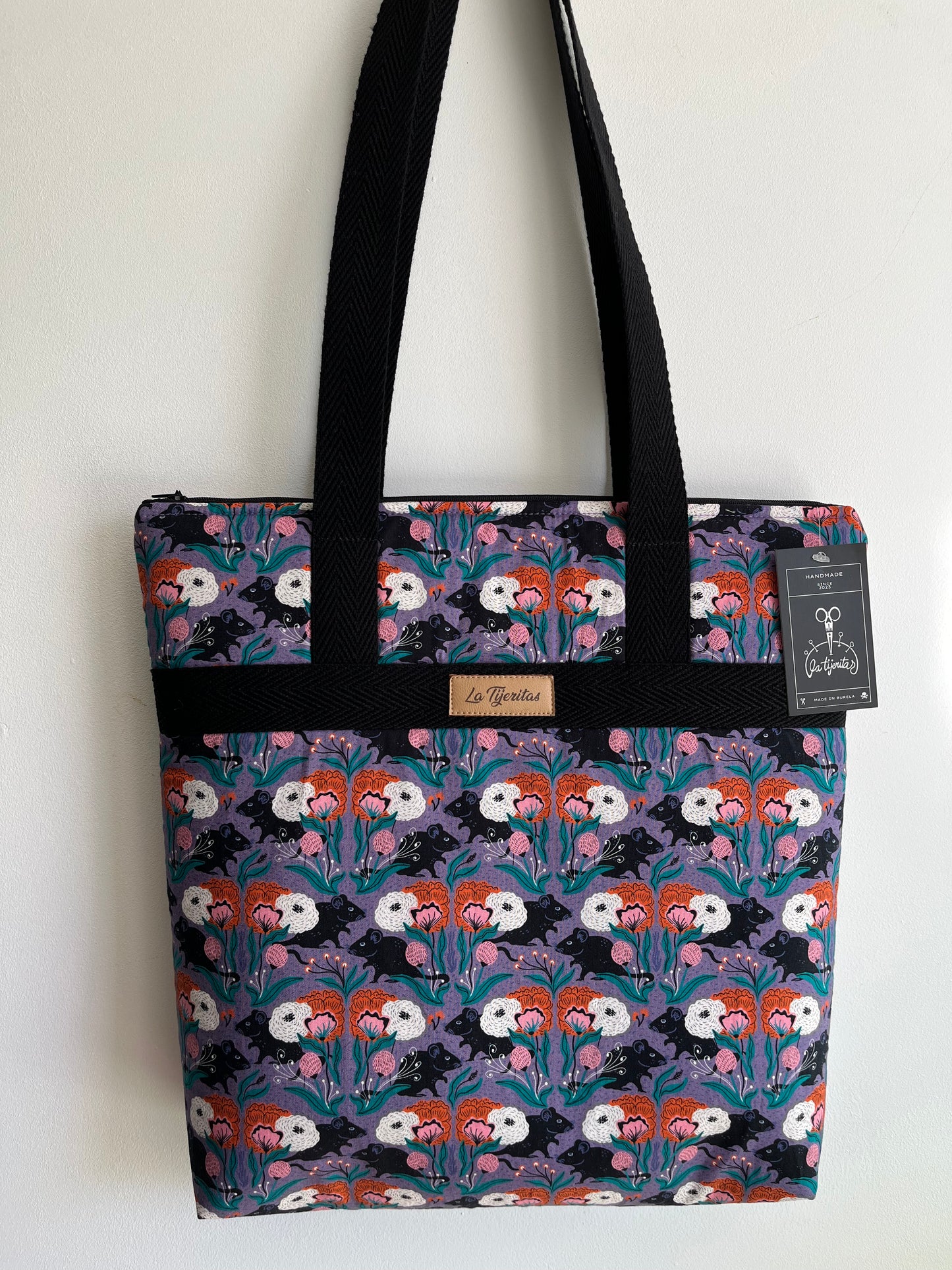 Bolso con estampado de ratitas negras y flores de color naranja y rosa. 
El fondo del estampado es de color lila.
Tiene tiras a contraste de color negro.